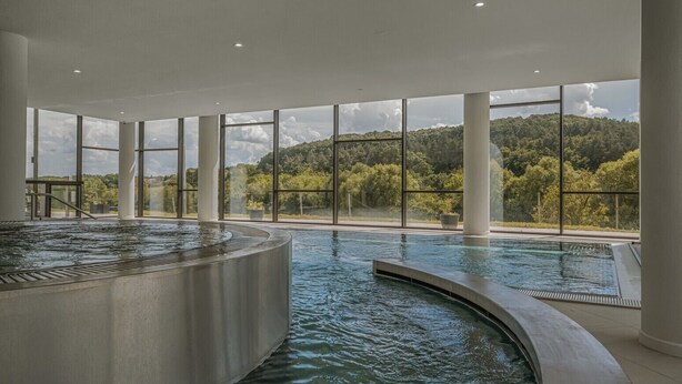 A modern indoor swimming pool with large windows offering a view of a forested hillside.