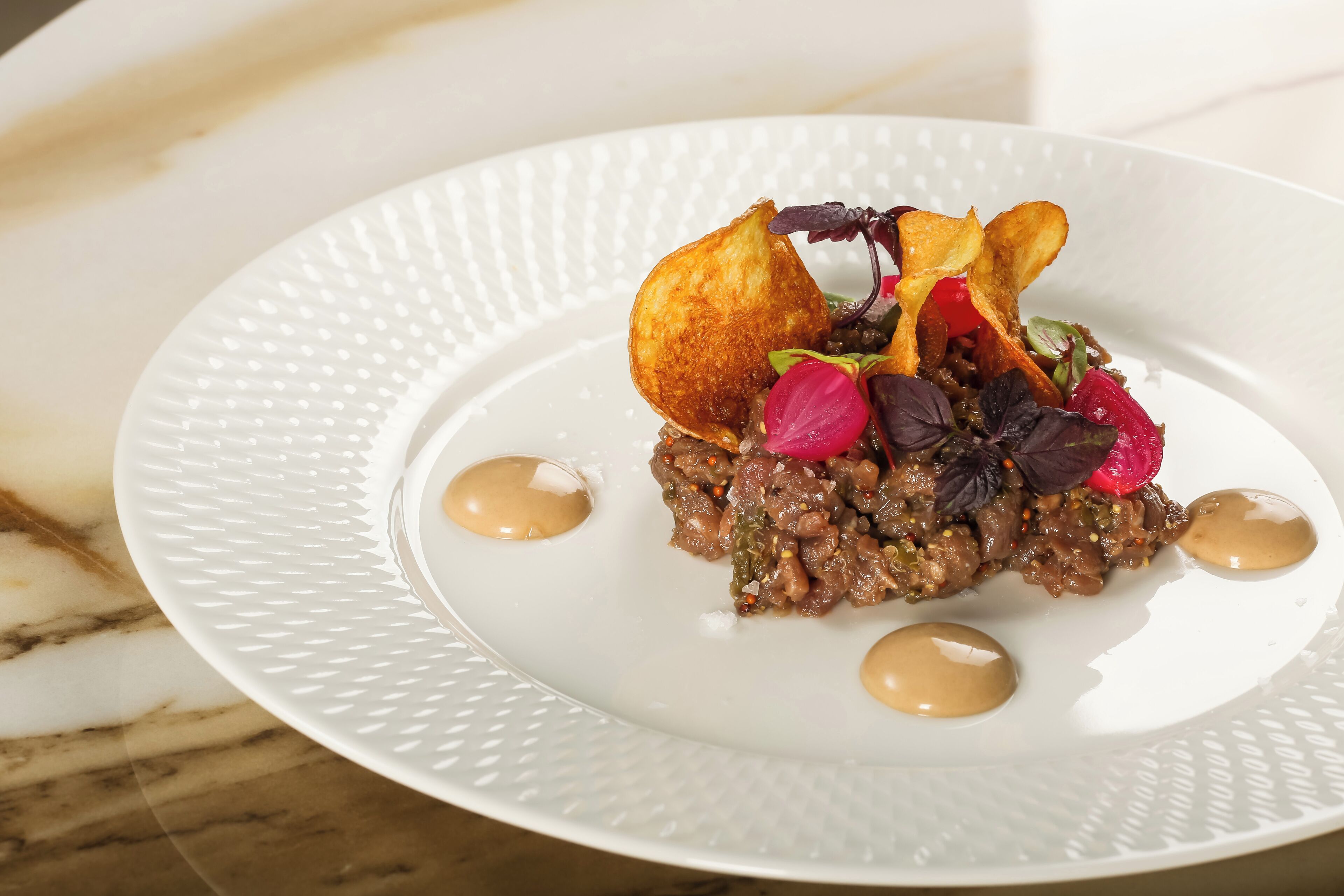 A white plate with a textured edge, containing a dish with a golden-brown item, green and red vegetables, and two round sauces.