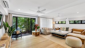 A modern living room with a sofa, ottoman, and a coffee table. There is a large sliding glass door leading to a balcony with plants. - Belle Escapes Sun Rose in Palm Cove (Palm Cove)