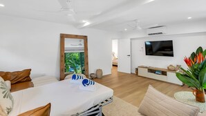 A modern living room with a flat-screen TV, a dining area with a table and chairs, and a view of greenery through a door. - Belle Escapes Sun Rose in Palm Cove (Palm Cove)