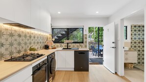 A modern kitchen with white cabinets, a built-in oven, and a dishwasher. There is a window with a view of the outdoors, and a door leading to a patio area. - Belle Escapes Sun Rose in Palm Cove (Palm Cove)