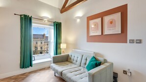 A modern living room with a sofa, a window with curtains, a radiator, and framed pictures on the wall. - Apartment with castle view (Alnwick)