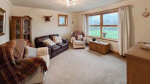 A cozy living room with a brown leather sofa, a plaid sofa, and a white armchair. A wooden cabinet and a small table with a vase of flowers are also visible.