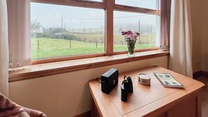 A room with a wooden desk, a vase with flowers, and a view of a green field through a window.