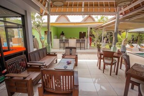 A covered outdoor seating area with wooden furniture, a bar, and a pool in the background. - Nanuk's Bungalows (Bali)