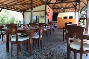 An outdoor dining area with wooden tables and chairs, a patterned floor, and a thatched roof structure. - Nanuk's Bungalows (Bali)