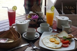 Een ontbijttafel met een bord eieren, toast en tomaten, een karaf sinaasappelsap, een theepot, kopjes en een mand.