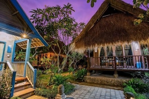 A thatched-roof building with a wooden deck, surrounded by tropical plants and a stone pathway. - Nanuk's Bungalows (Bali)