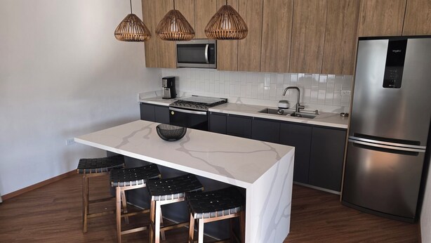 Een moderne keuken met een wit kookeiland, zwarte barkrukken, een roestvrijstalen koelkast en houten keukenkastjes.