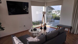Een moderne woonkamer met een grijze bank, een ronde salontafel en een groot raam met uitzicht op een stadsgezicht.