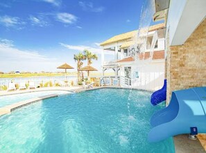 Una piscina con tobogán de agua, palmeras y un edificio con techo de paja al fondo.
