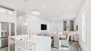 A modern kitchen with white cabinetry, a large island, and stainless steel appliances. There is a dining area with a white table and chairs, and a living room with a sofa and a TV. - Luxury Home | Golf Cart | Wavecrest Retreat | Pool | Inlet Beach (Inlet Beach)