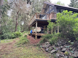 A wooden deck with a person working on it, surrounded by trees and a rocky terrain. - Cozy, secluded 3 bedroom ,2 bathroom home. Where the sea and forest meet (Sooke)
