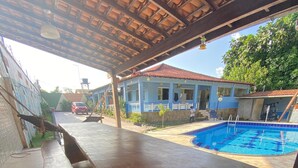 A covered outdoor area with a pool, wooden furniture, and a building in the background.