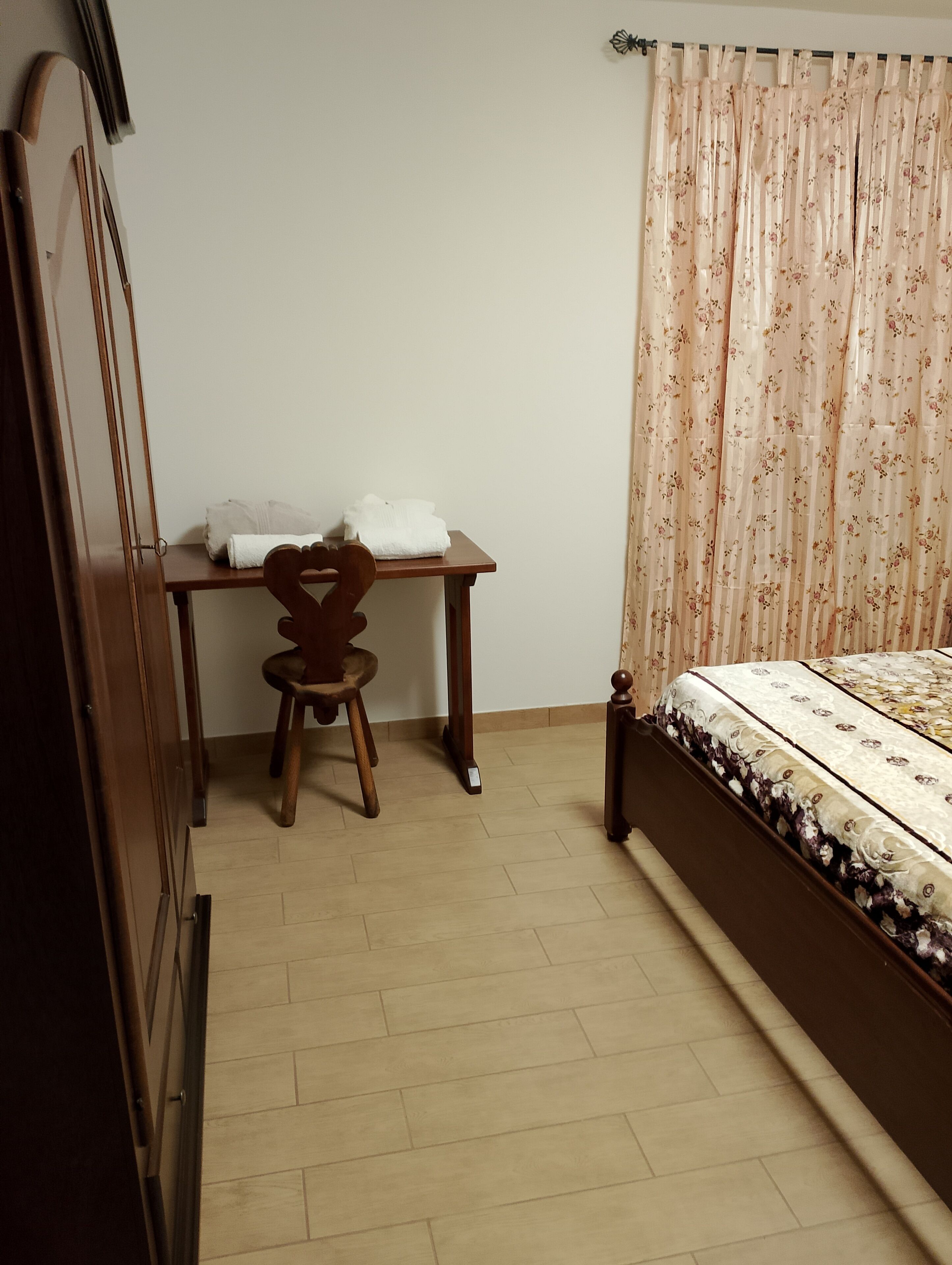 Ein Schlafzimmer mit einem hölzernen Bett, einem Schreibtisch mit Stuhl und einem Fenster mit blumigen Vorhängen.