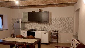 Une cuisine compacte avec des meubles blancs, une cuisinière à gaz et un évier. On y trouve une table à manger avec des chaises et un fauteuil à motifs.