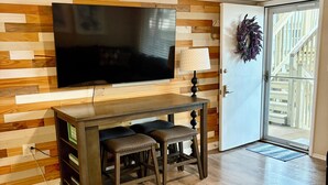 Un salon doté d’un téléviseur à écran plat, de lambris en bois, d’une porte avec un panneau de verre et d’un paillasson bleu.