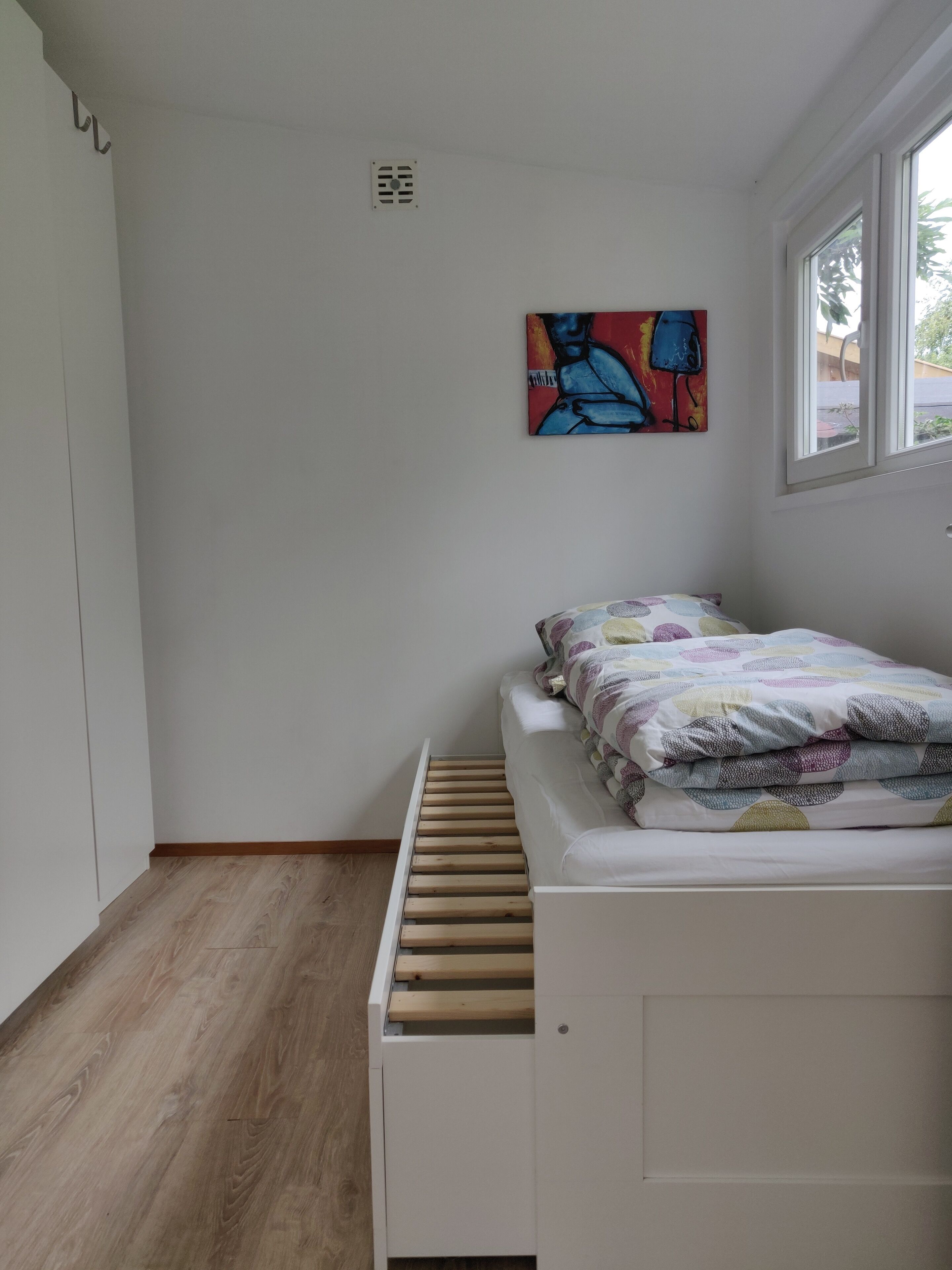 Une chambre à coucher avec un lit, une fenêtre, une œuvre d’art accrochée au mur et un plancher en bois.