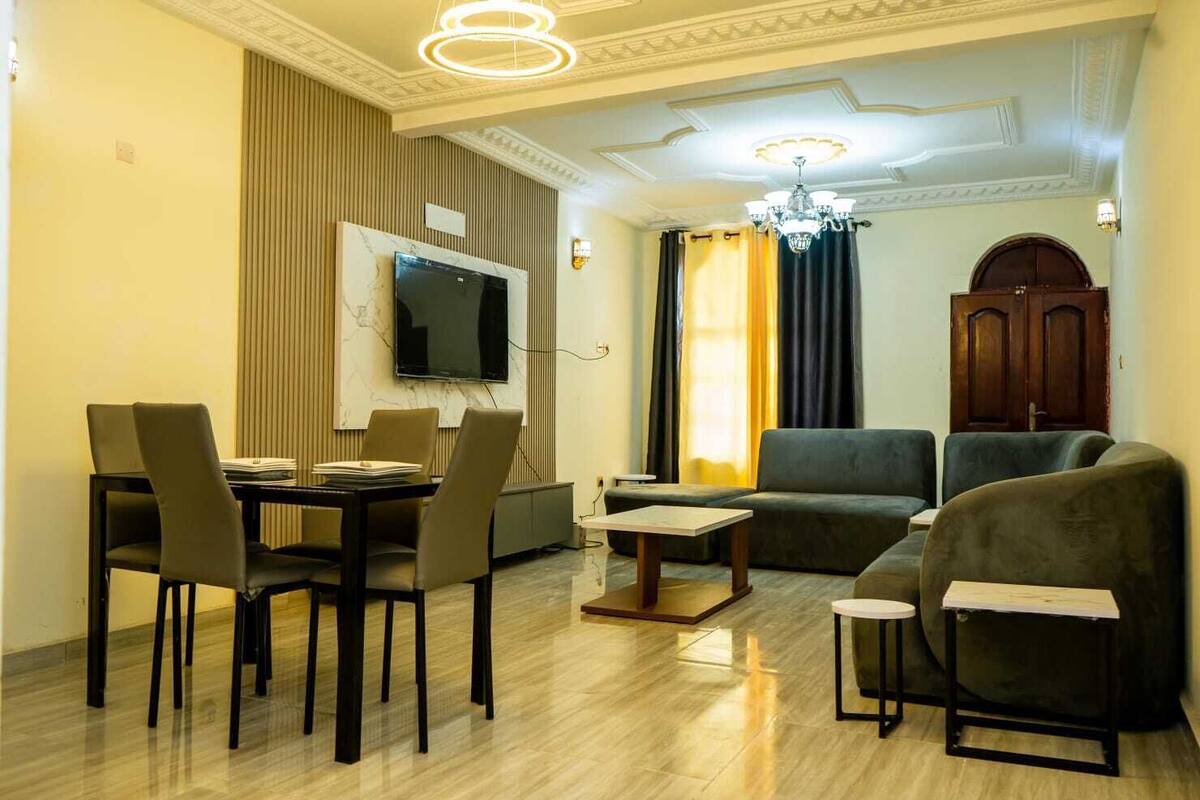 A modern living room with a dining area, a flat-screen TV mounted on the wall, and a chandelier.