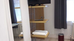 A compact living space with a television mounted on a wooden shelf, a small white table, a red trash bin, and a patterned rug on the floor. - Number Four  (Brighton)