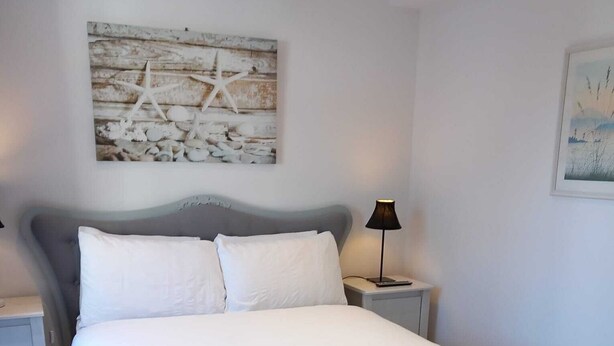 A neatly made bed with white linens and two folded towels, a nightstand with a lamp, and a rustic wall art piece above the bed. - Number Four  (Brighton)