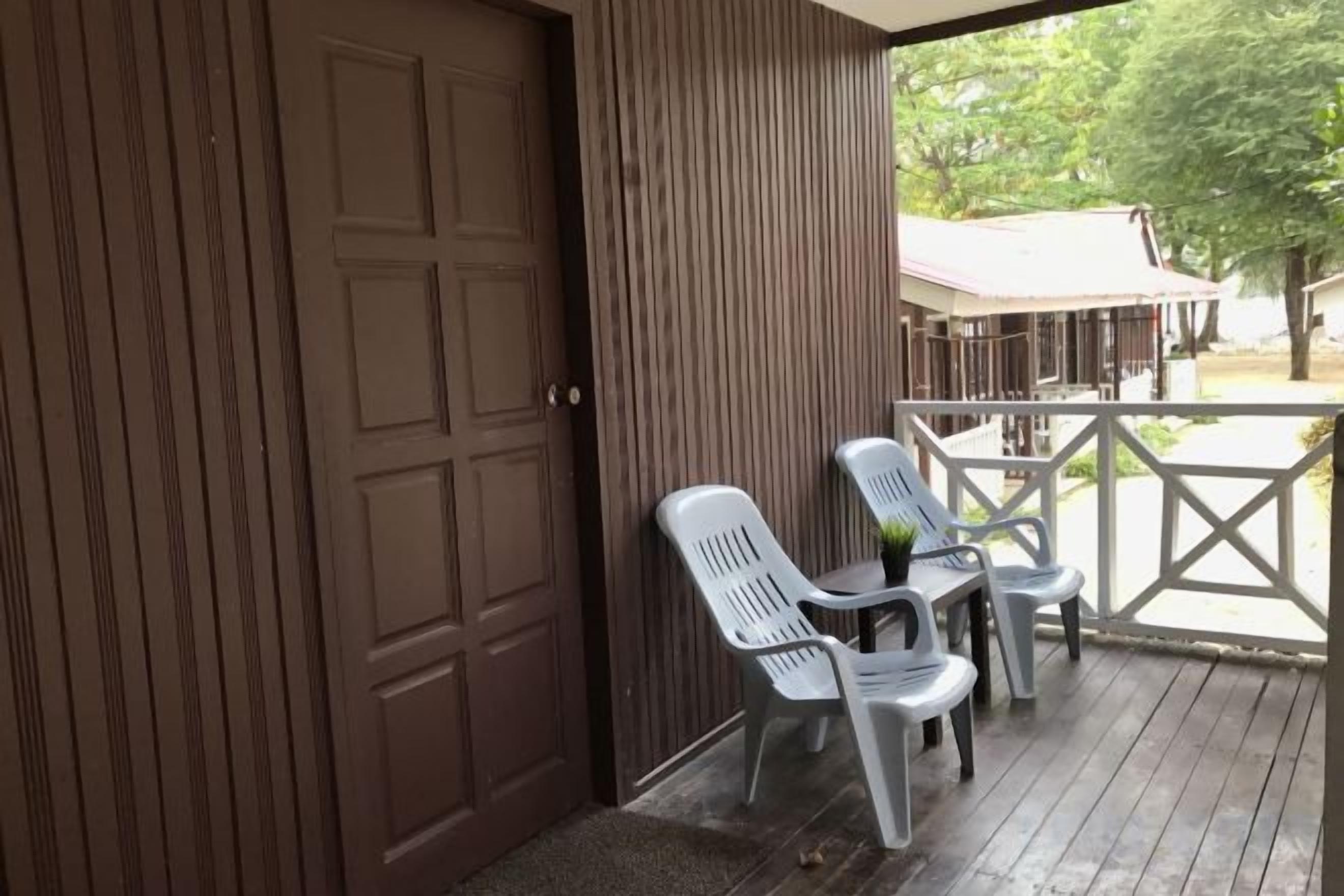 A covered porch with a wooden door and a railing. There are two white plastic chairs and a small table.