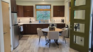 Una cocina moderna con armarios de madera, un refrigerador de acero inoxidable, una mesa de comedor con sillas y un amplio ventanal con vista a árboles.