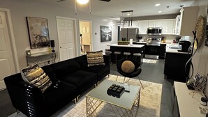 A modern living room with a black sofa, a coffee table, and a round ottoman. There is a kitchen in the background with white cabinets and a microwave. - Double Up In Delano-by Dtwn Stadium&ballpark Arena (Wichita)