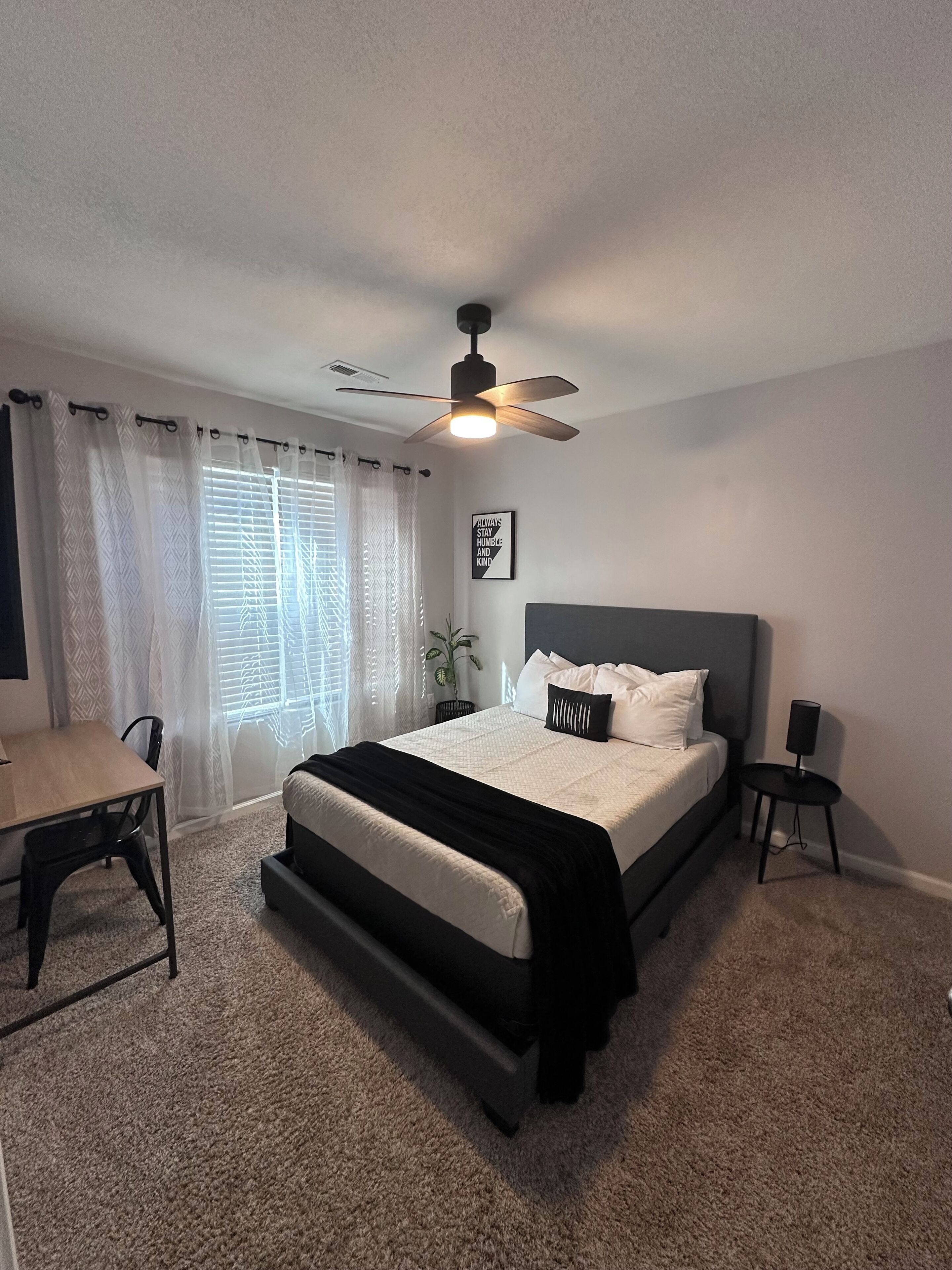 Un dormitorio con cama, escritorio, silla, ventilador de techo y ventana con cortinas.