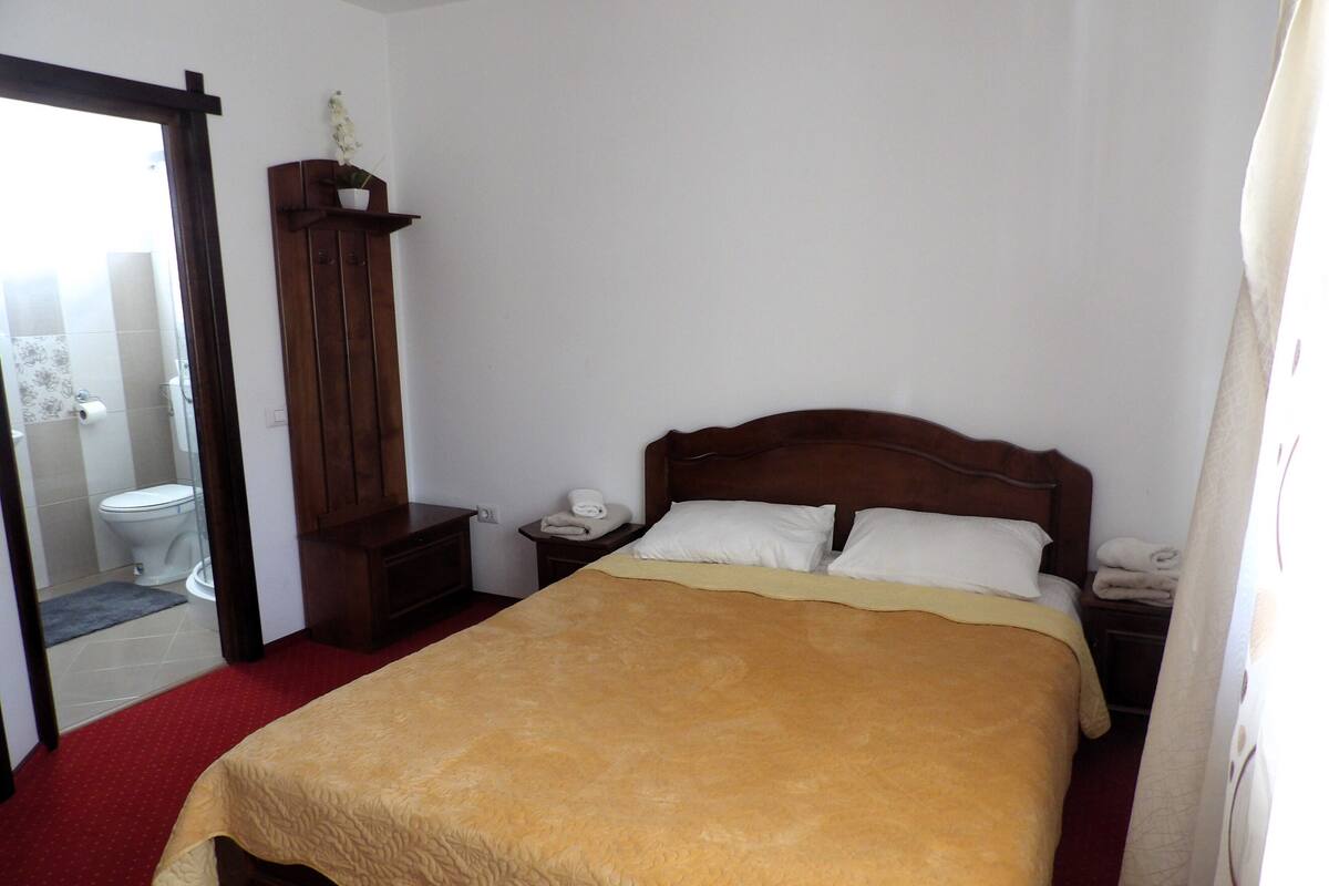A bedroom with a large bed, a wooden nightstand, a bedside table, a wooden wardrobe, and a bathroom visible through an open door.