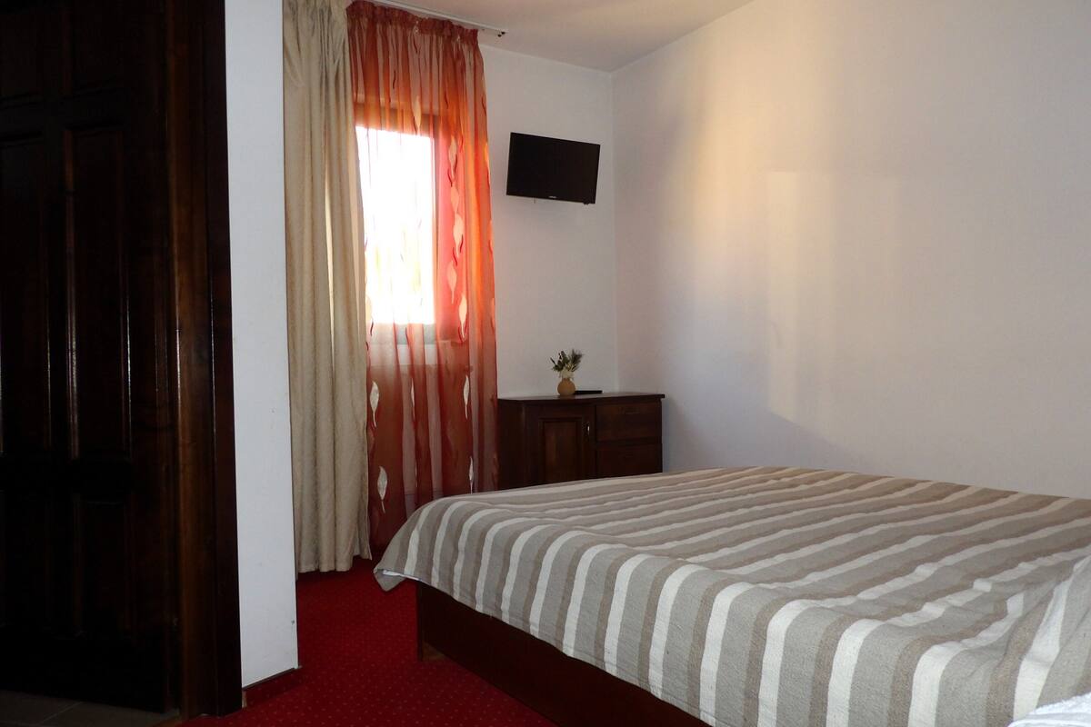A bedroom with a bed, a TV mounted on the wall, a wooden wardrobe, and a window with curtains.