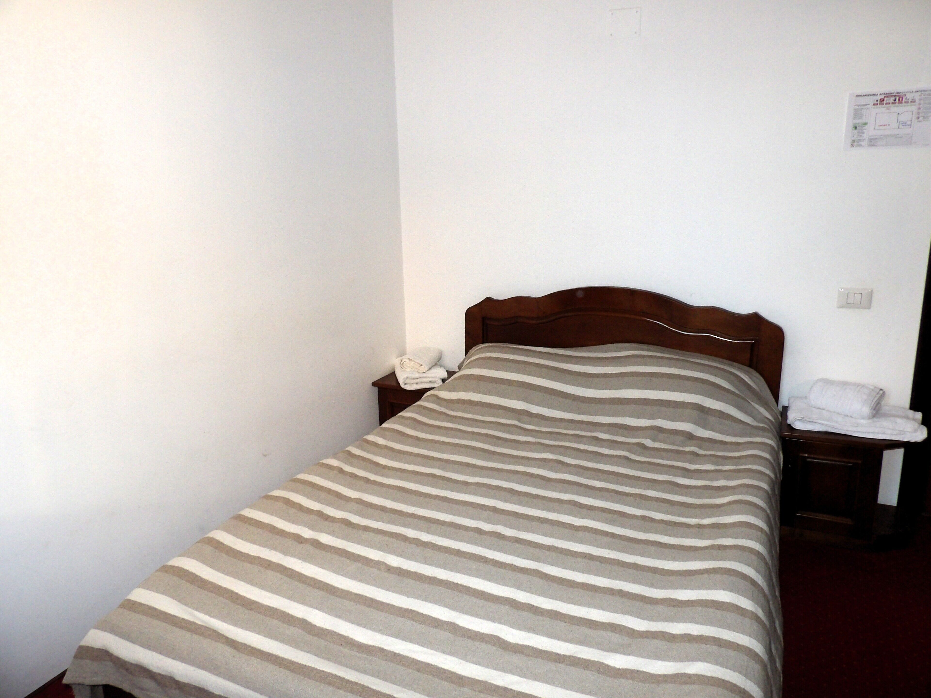 A single bed with a striped bedspread, a wooden headboard, a nightstand with a telephone, and a wall-mounted light switch.