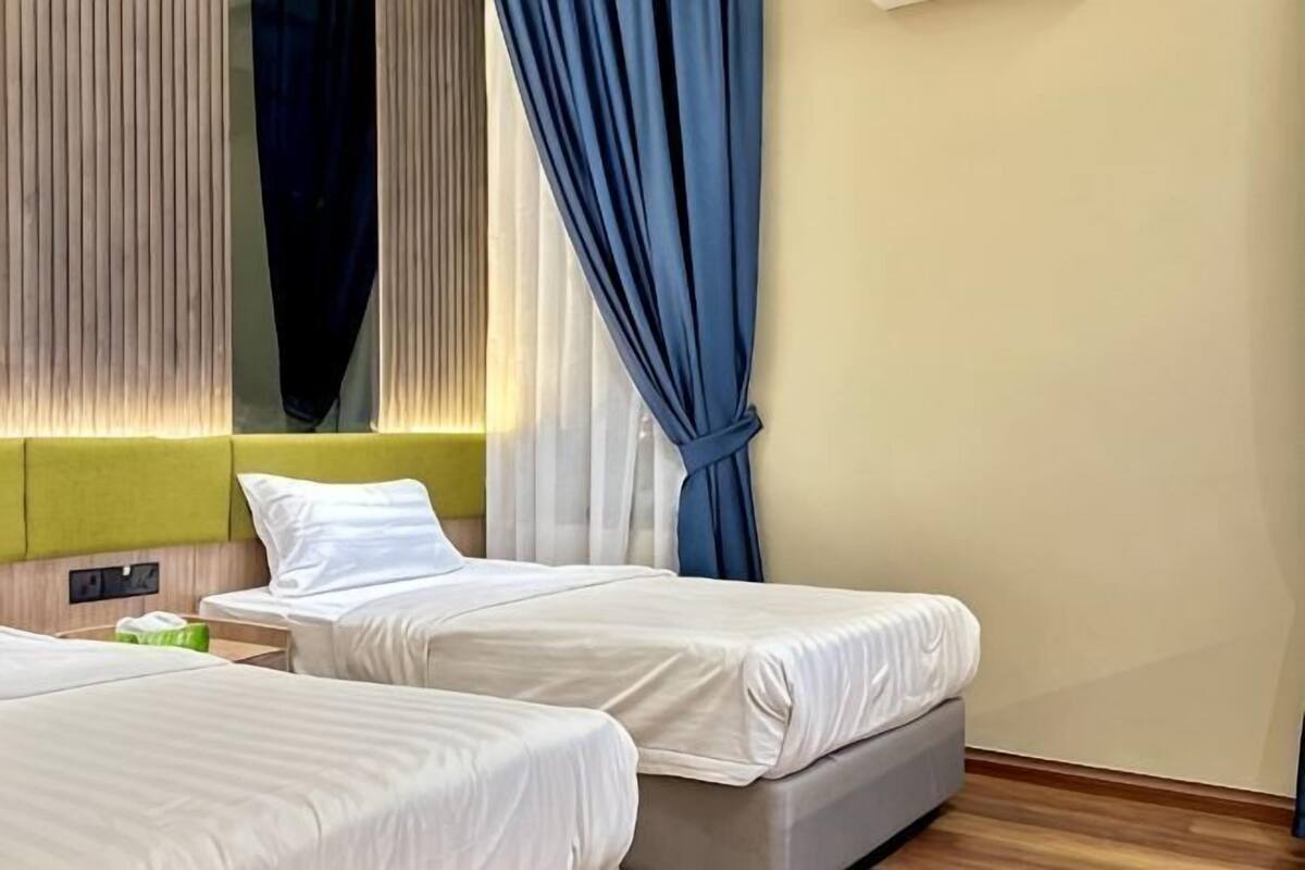 A hotel room with two beds, a green headboard, a bedside table, and a window with blue curtains.