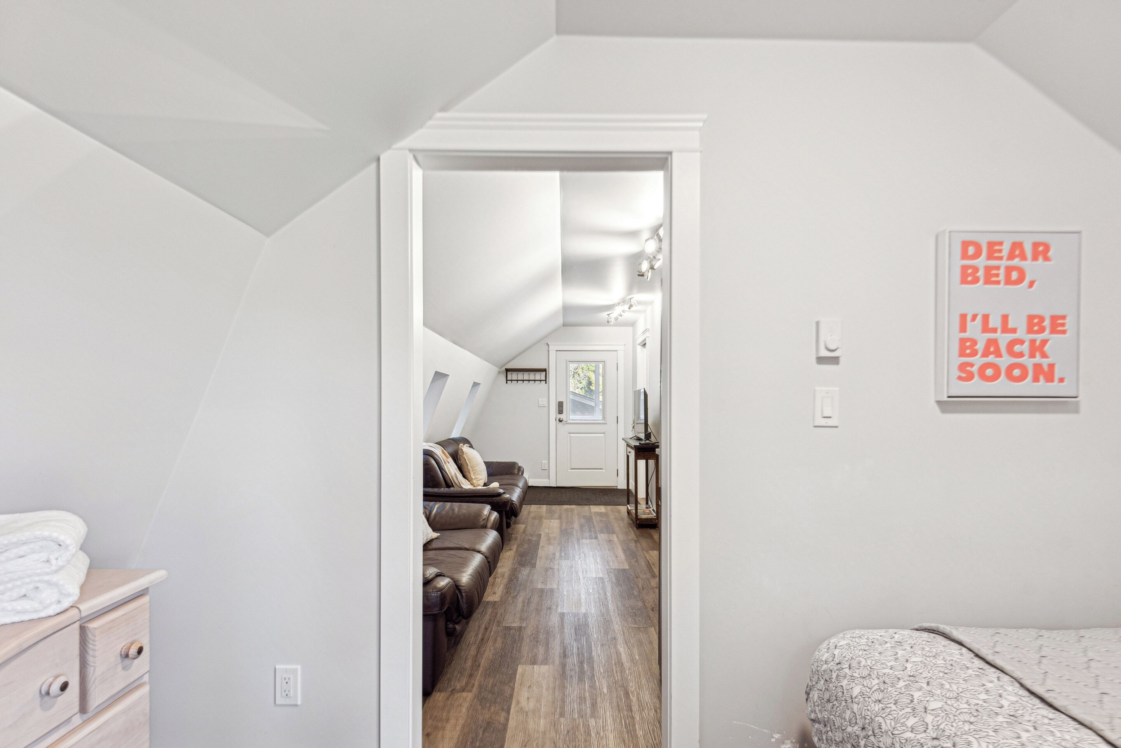 Un couloir étroit mène à une chambre où se trouvent un lit et une chaise. Une pancarte sur le mur porte l’inscription : « CHER LIT, JE REVIENDRAI BIENTÔT. »