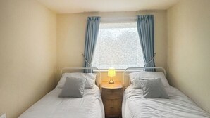 Un petit logement composé d’une seule pièce, comprenant deux lits simples, une table de chevet avec une lampe et une fenêtre avec des rideaux.