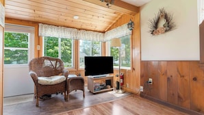Una habitación acogedora con techo de madera, una silla de mimbre, un televisor de pantalla plana y ventanas amplias con cortinas.