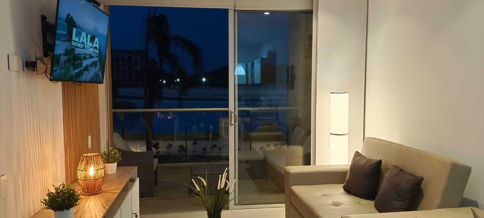 A modern living room with a sofa, a TV mounted on the wall, a glass door leading to an outdoor area, and a small table with a lamp and plants.