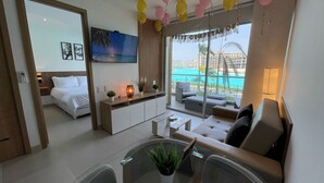 Habitación de hotel moderna con cama, sofá, zona de comedor y vistas a una piscina y playa.