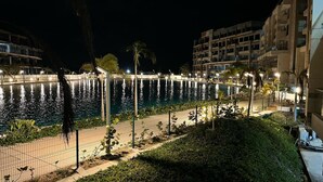 Vista nocturna de un paseo marítimo con piscina, palmeras y edificios al fondo.