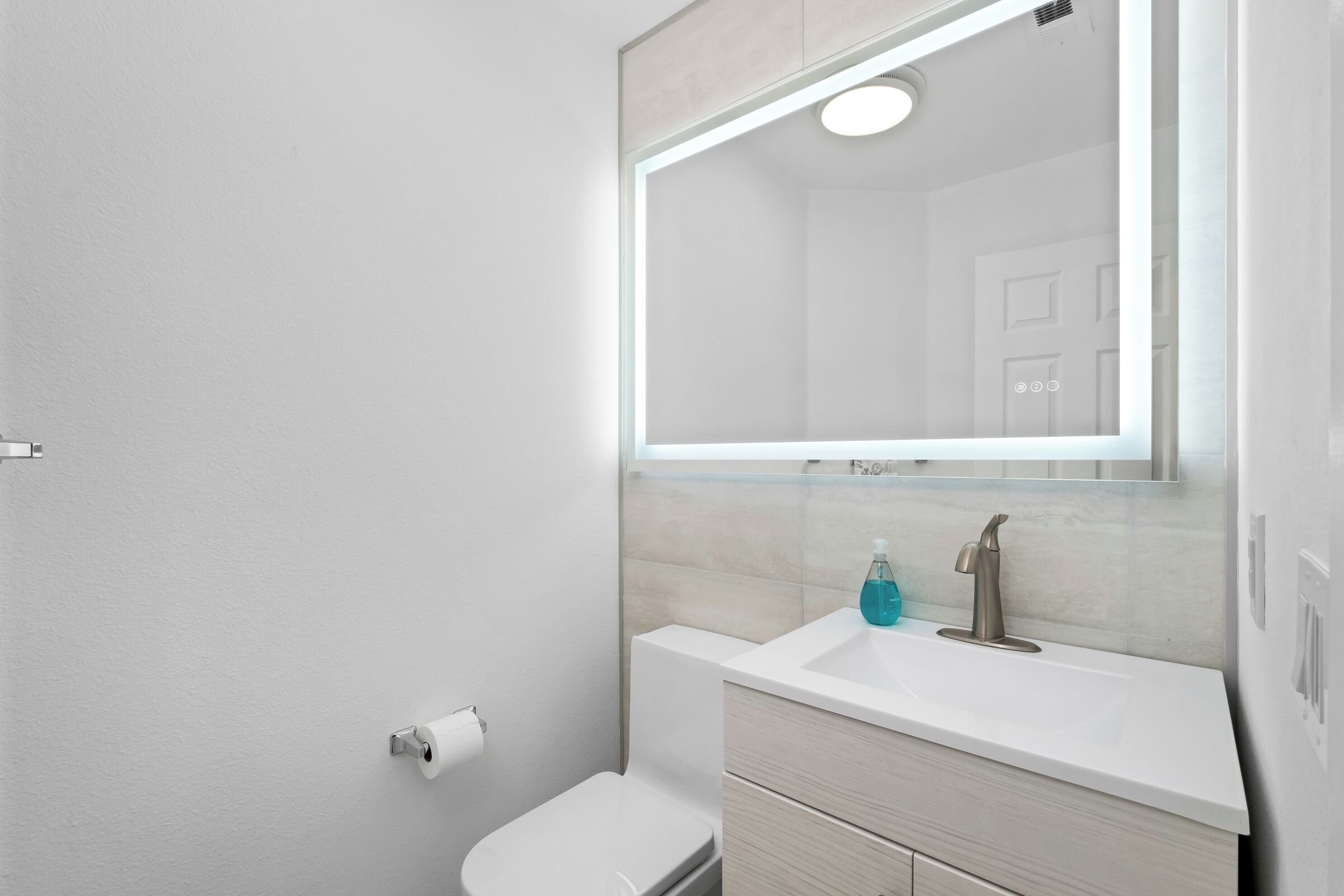 A modern bathroom with a white toilet, a sink with a mirror above it, and a blue soap dispenser.