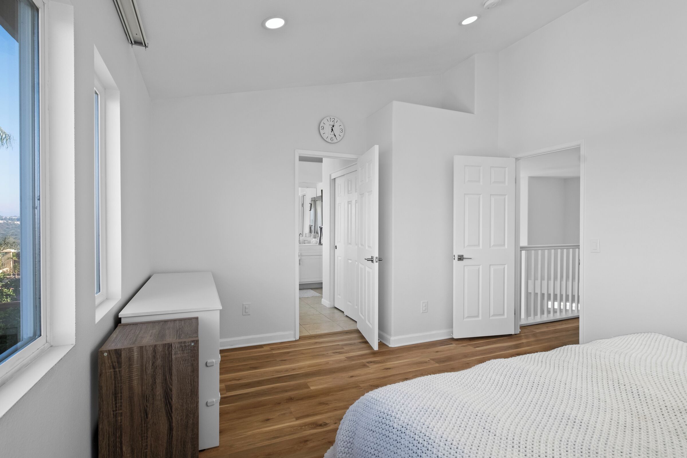 A modern bedroom with a large window, wooden flooring, a white bed, and a small desk.