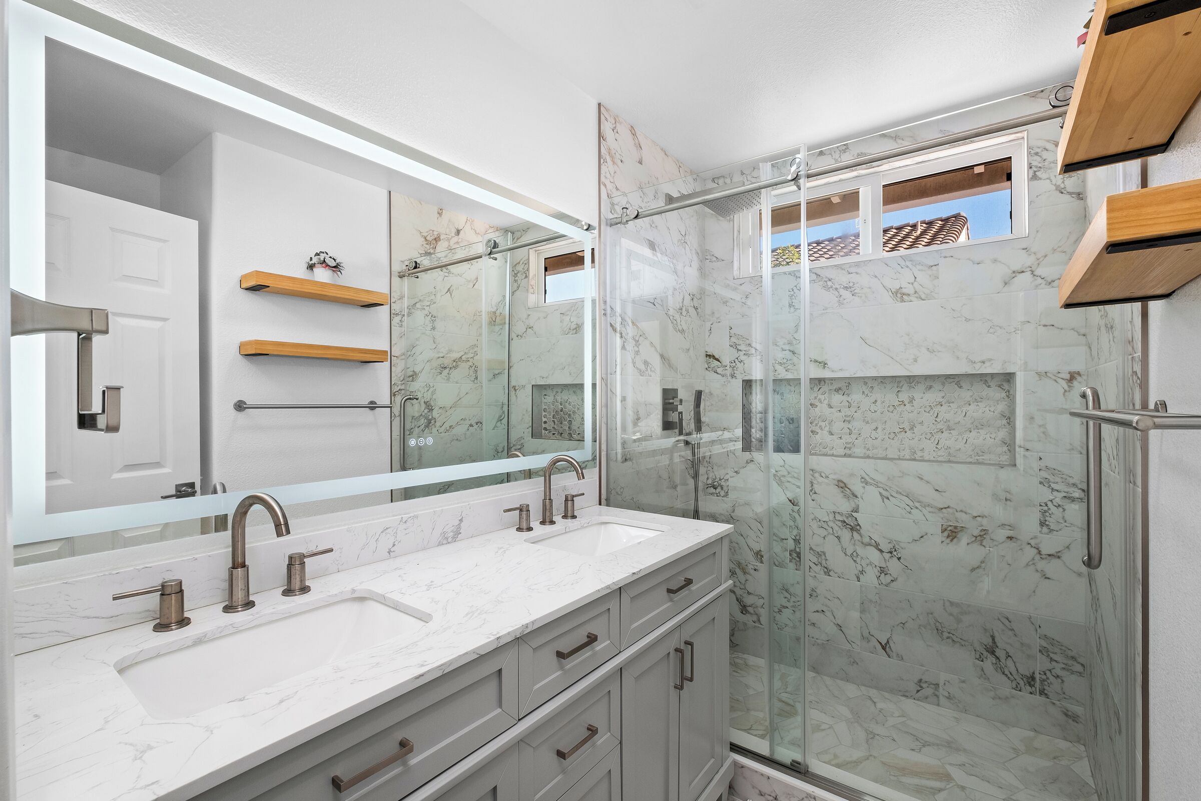 A modern bathroom with a large mirror, two sinks, a shower enclosure, and marble walls.