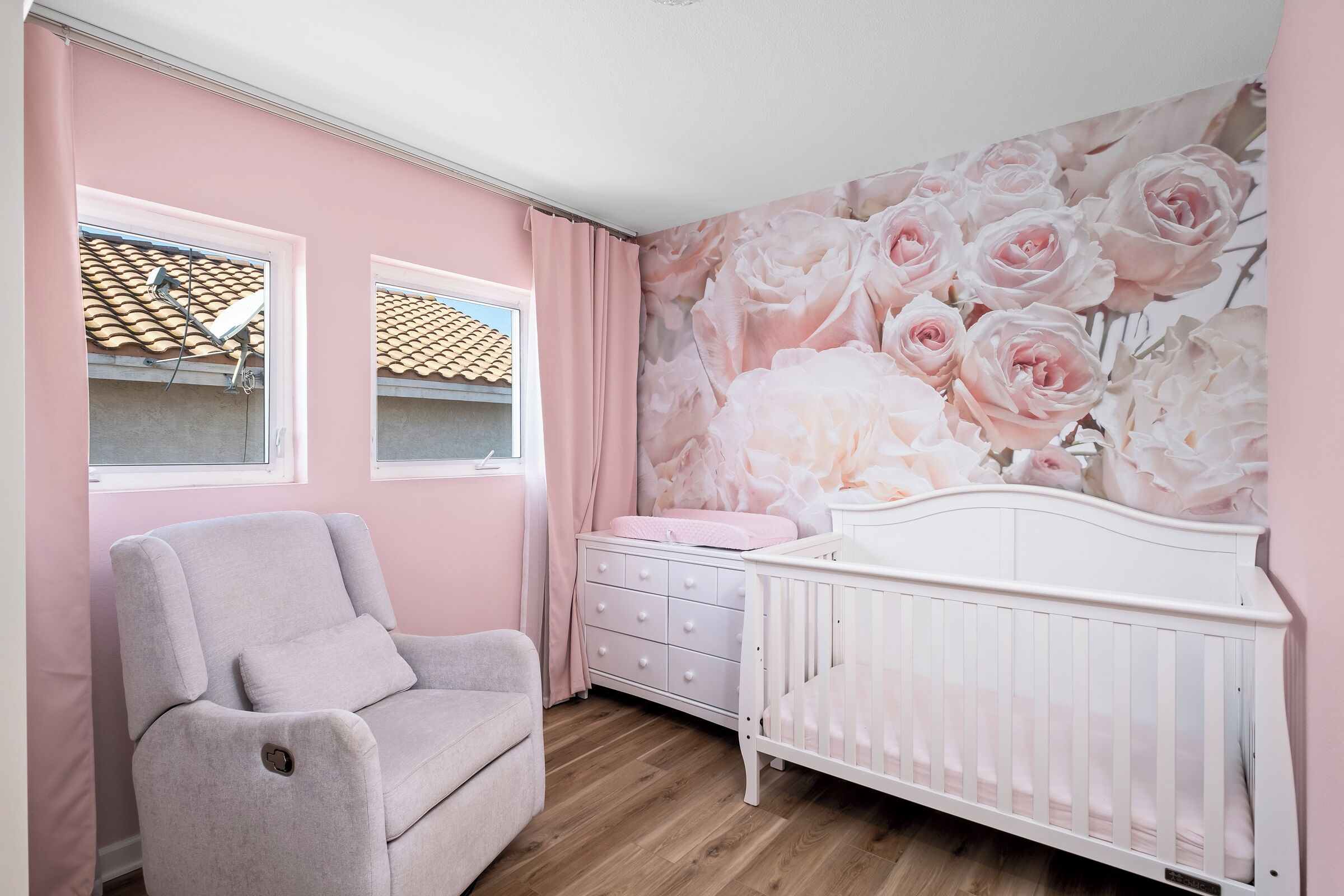 Une pièce aux murs roses, avec un berceau blanc, une chaise grise et une commode.