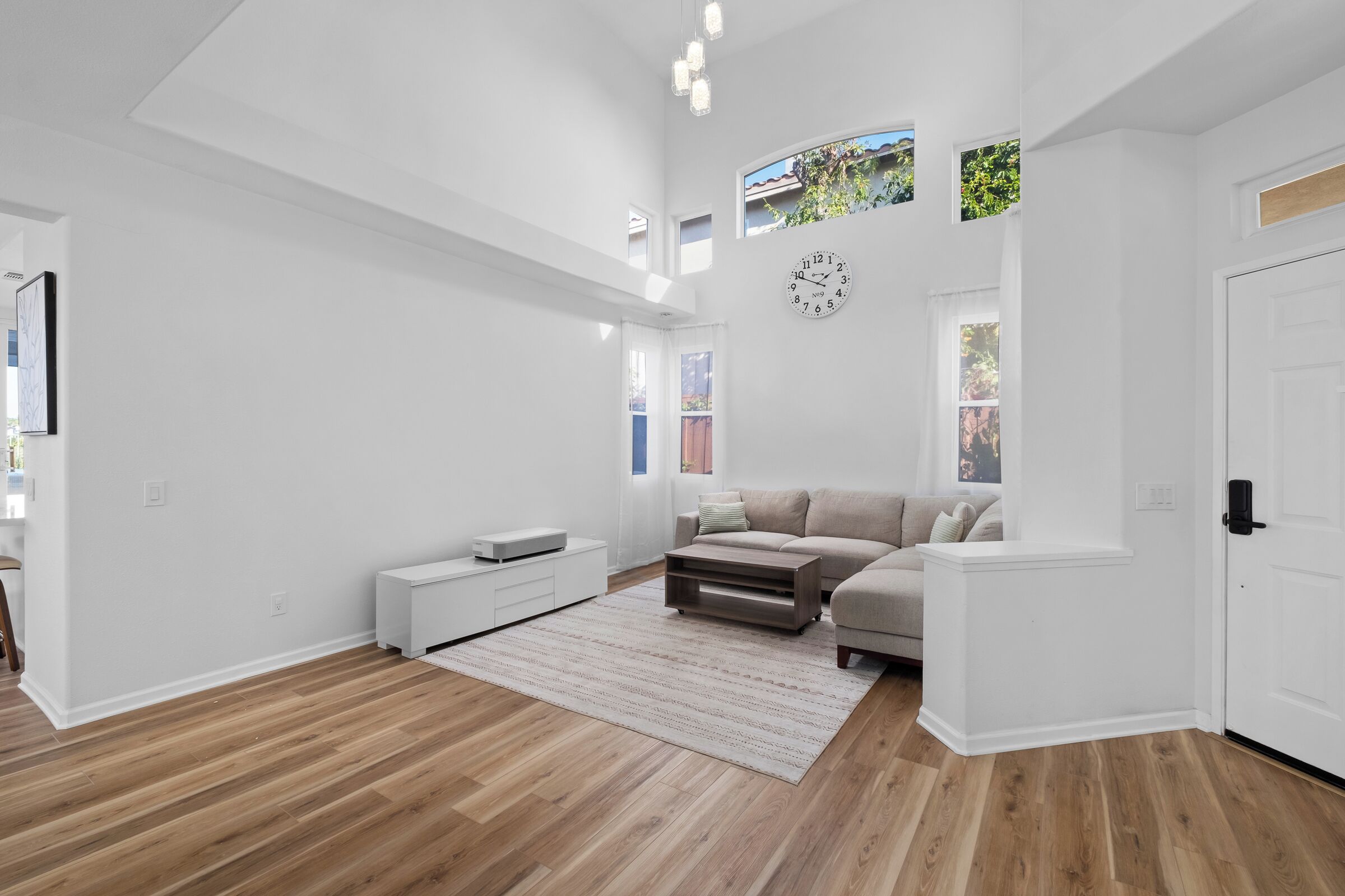 Een ruime woonkamer met een houten vloer, een beige bank, een witte salontafel en een grote klok aan de muur.