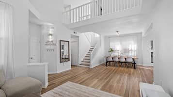 A spacious living area with a staircase leading to an upper floor, a dining area with a table and chairs, and a large window providing natural light.