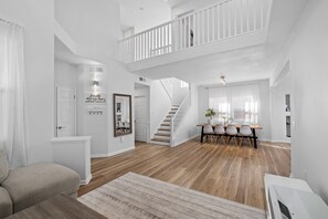 Een ruime woonkamer met een trap naar een verdieping hoger, een eethoek met een tafel en stoelen, en een groot raam voor natuurlijk licht.