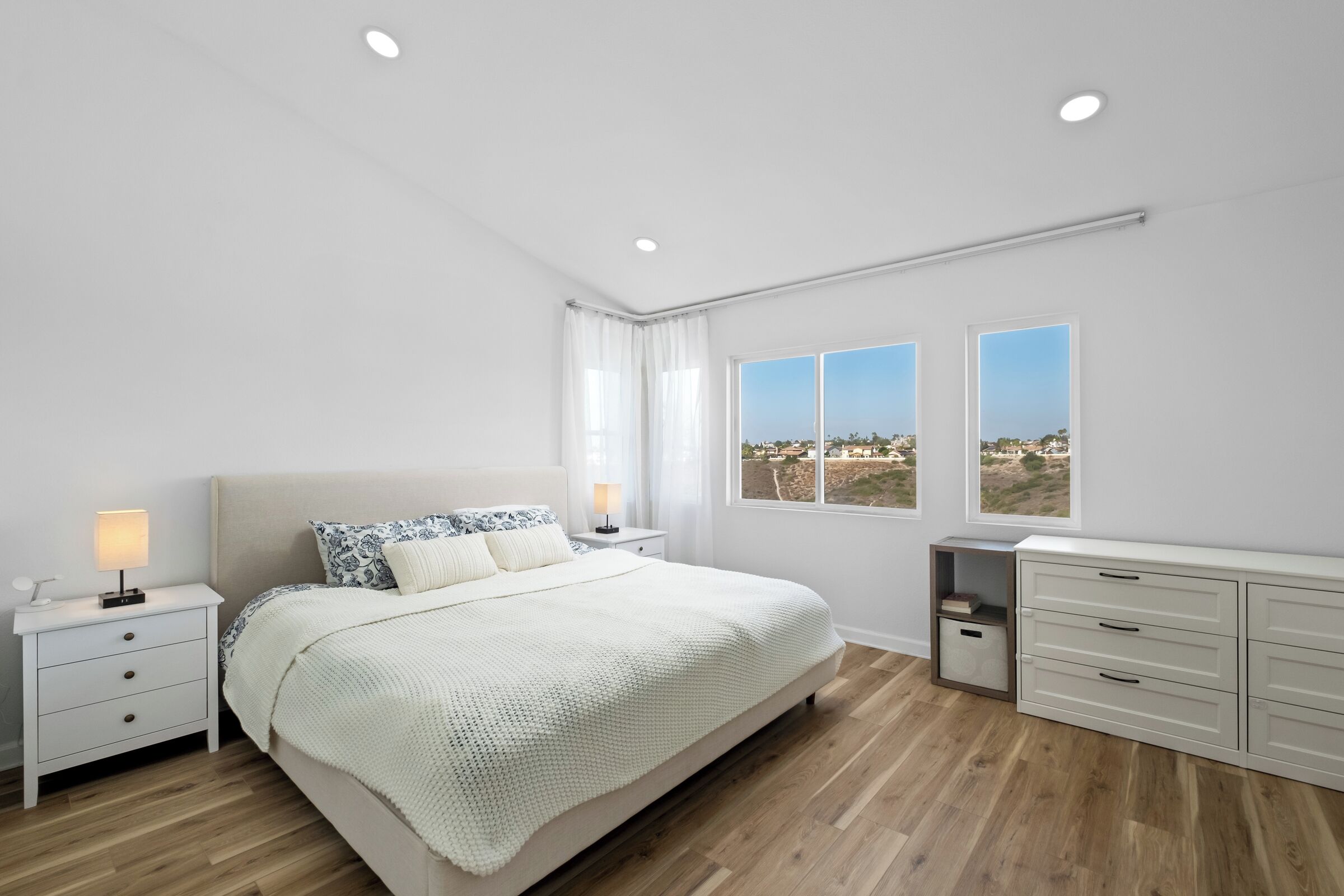 A modern bedroom with a large bed, bedside tables, a dresser, and two windows offering a view.