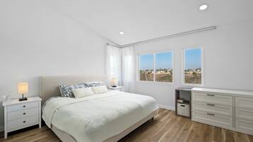 A modern bedroom with a large bed, bedside tables, a dresser, and two windows offering a view.
