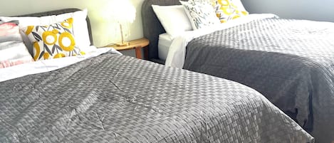 A hotel room with two beds, each with grey bedding and patterned pillows. There is a small bedside table with a lamp, and the walls are painted light blue.