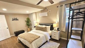 A bedroom with a bed, bedside tables, a ceiling fan, an air conditioning unit, a wall-mounted shelf, and a door.